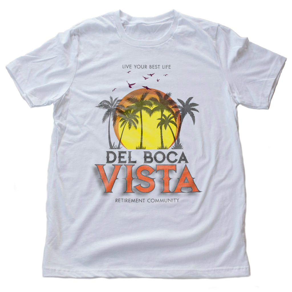 A retro-styled, colorful graphic t-shirt, referencing the many Seinfeld tv episodes featuring Del Boca Vista, a fictional Florida retirement home. This shirt features palm trees and birds against a setting sun, and the words "Del Boca Vista Retirement Community" beneath.