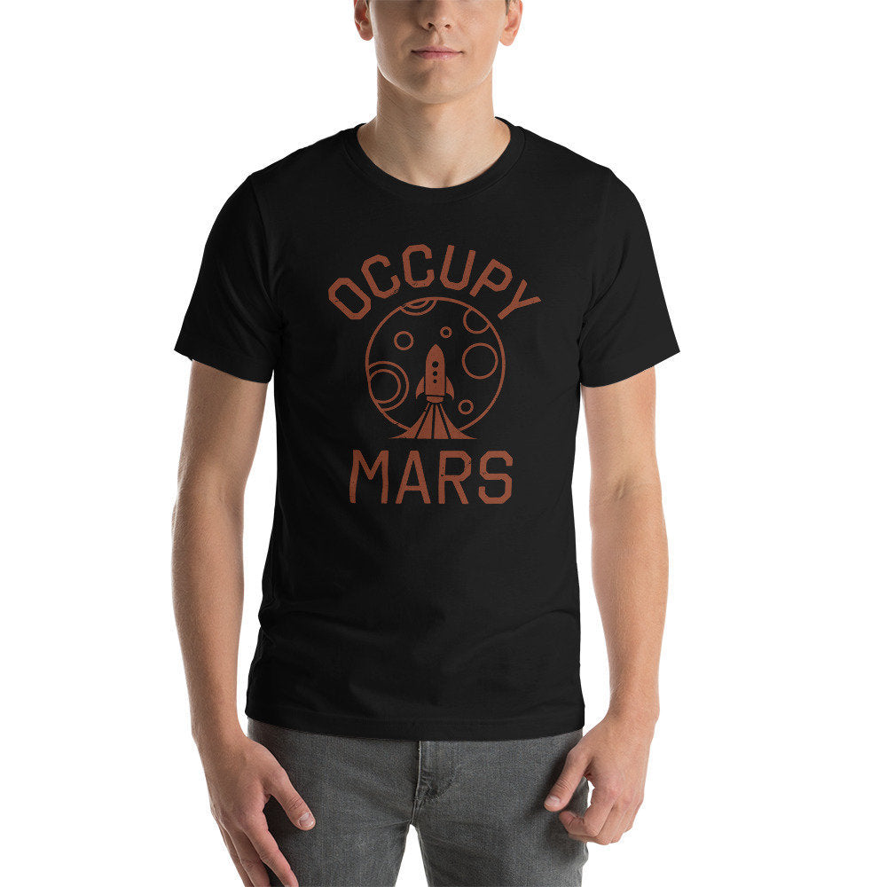 Male model wearing a sarcastic, classic, retro-inspired graphic t-shirt featuring the illustration of a rocketship blasting off toward a planet, and the meme words "OCCUPY MARS" encircling it, perhaps mocking Elon Musk and SpaceX.