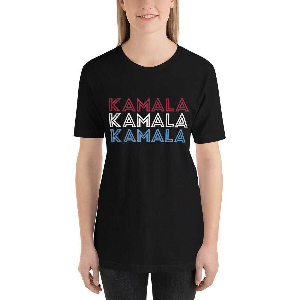 Female model wearing a classic graphic t-shirt featuring the Vice President's name three times in a vertical stack, in red, white, and blue: "KAMALA KAMALA KAMALA" celebrating her win with Joe Biden over Donald Trump in November 2020.