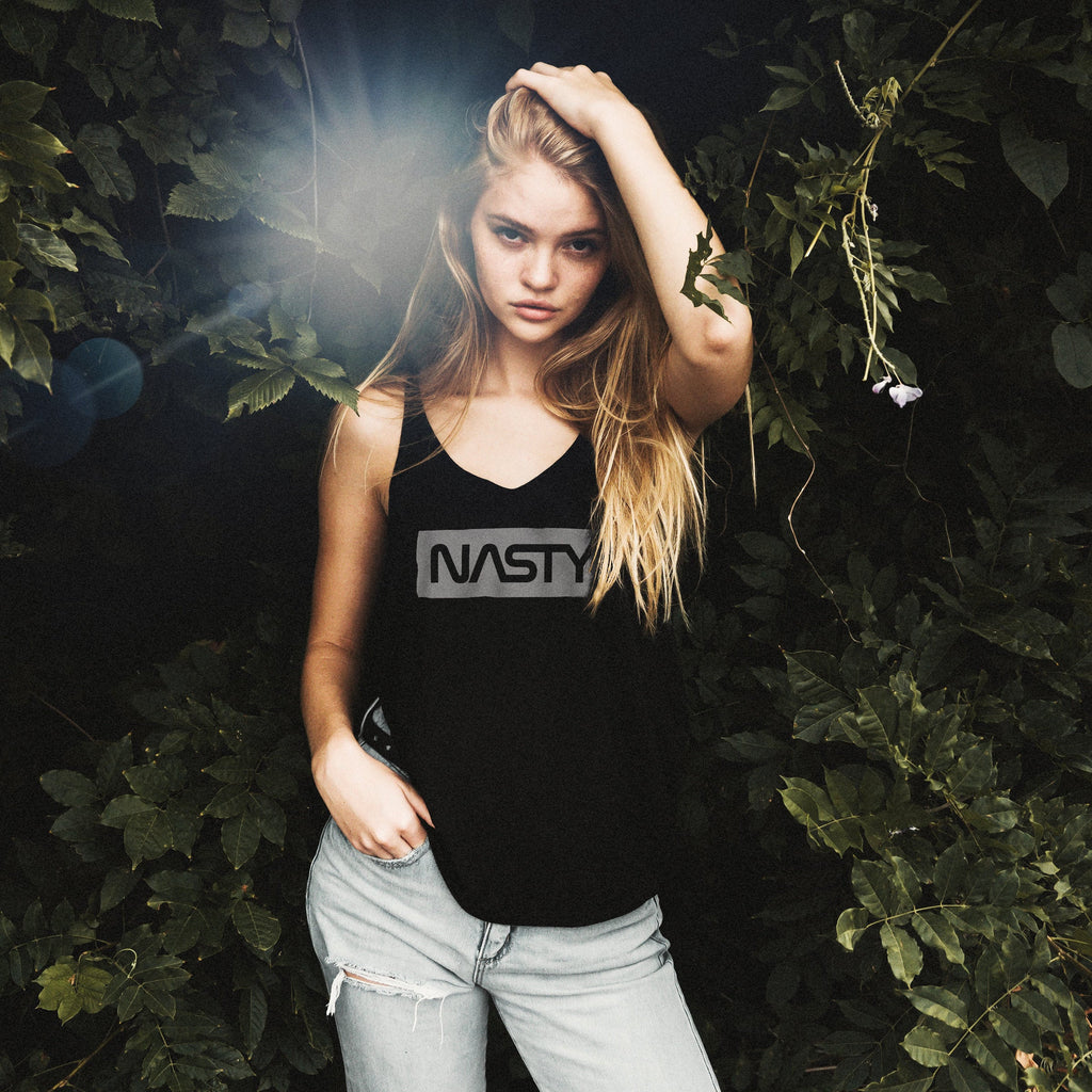 Model against the backdrop of green-leaved trees, wearing a fashionable graphic t-shirt featuring a parody of the NASA 'noodle' logo, reading "NASTY" instead of NASA.